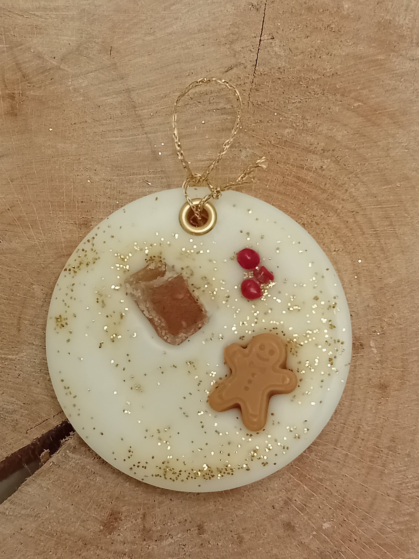 Circular wax sachet decorated with a piece of crystallized ginger, a wax gingerbread man, pink peppercorns and gold glitter with a wooden log slice background.
