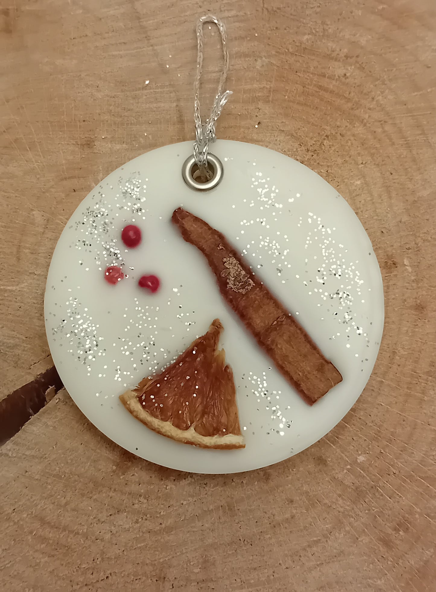 Circular wax sachet decorated with a cinnamon stick, orange slice, pink peppercorns and silver glitter with a wooden log slice background.