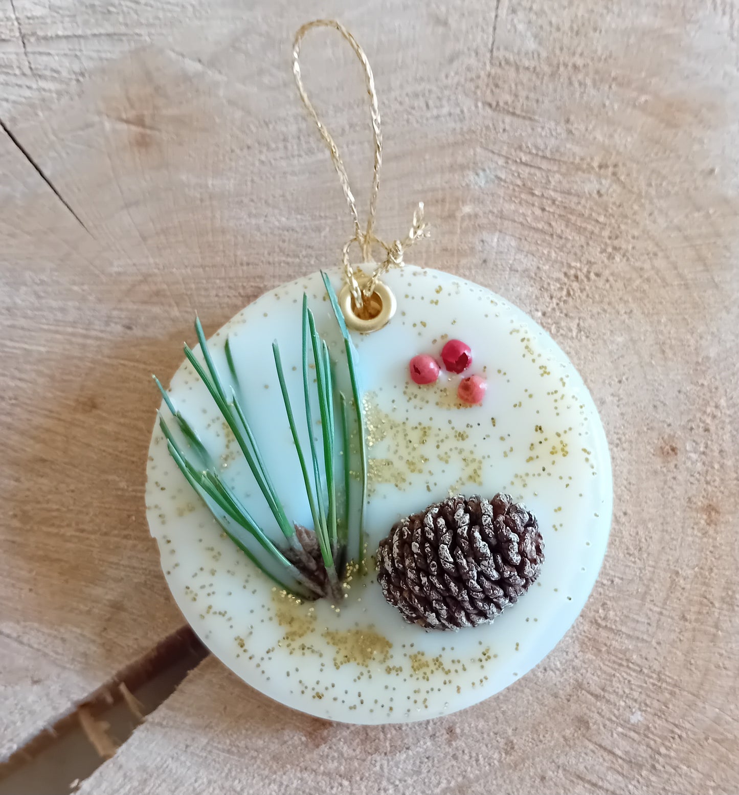 Circular wax sachet decorated with a birch pine cone, pine tree leaves, pink peppercorns and gold glitter with a wooden log slice background.
