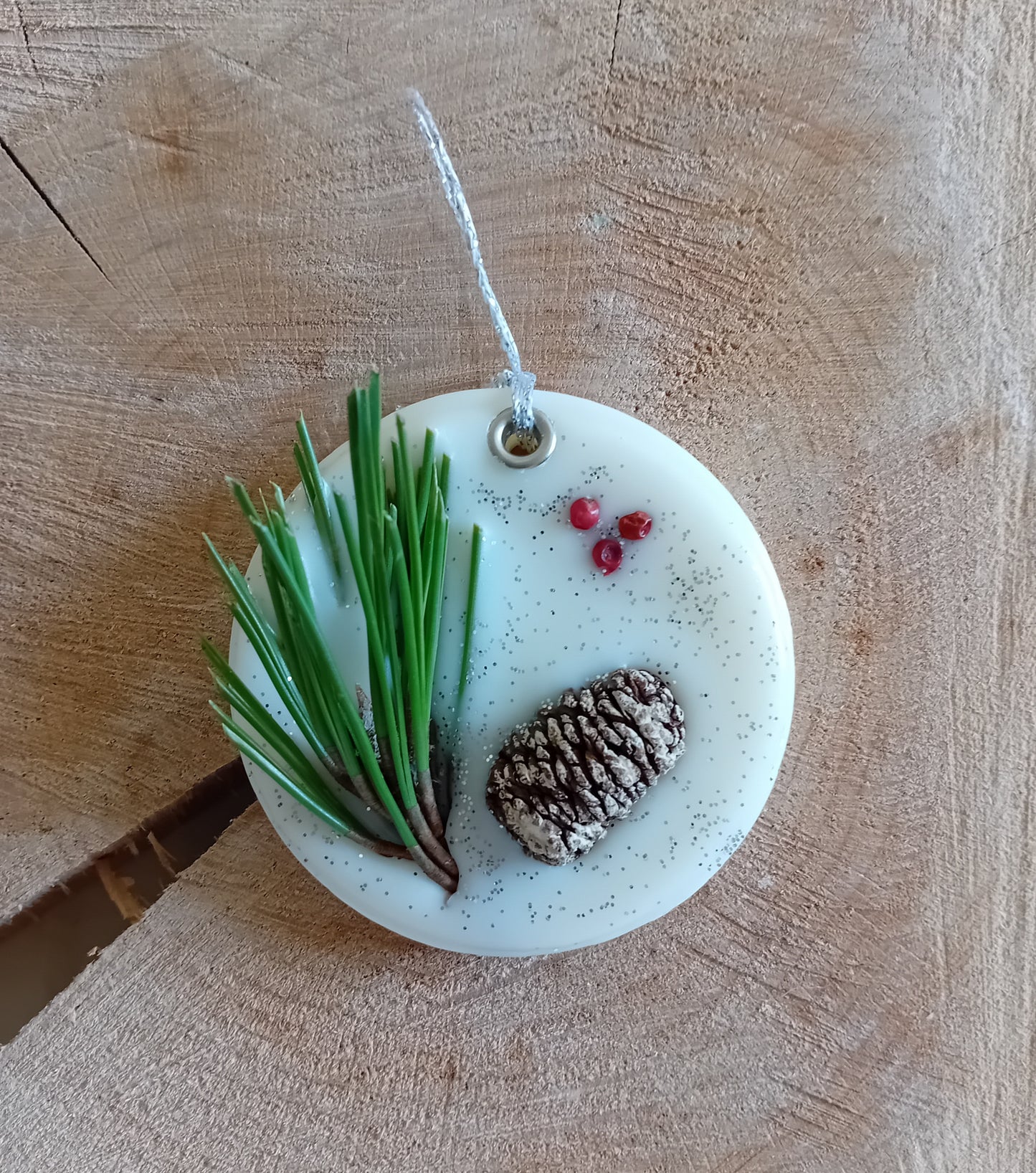 Circular wax sachet decorated with a birch pine cone, pine tree leaves, pink peppercorns and silver glitter with a wooden log slice background.