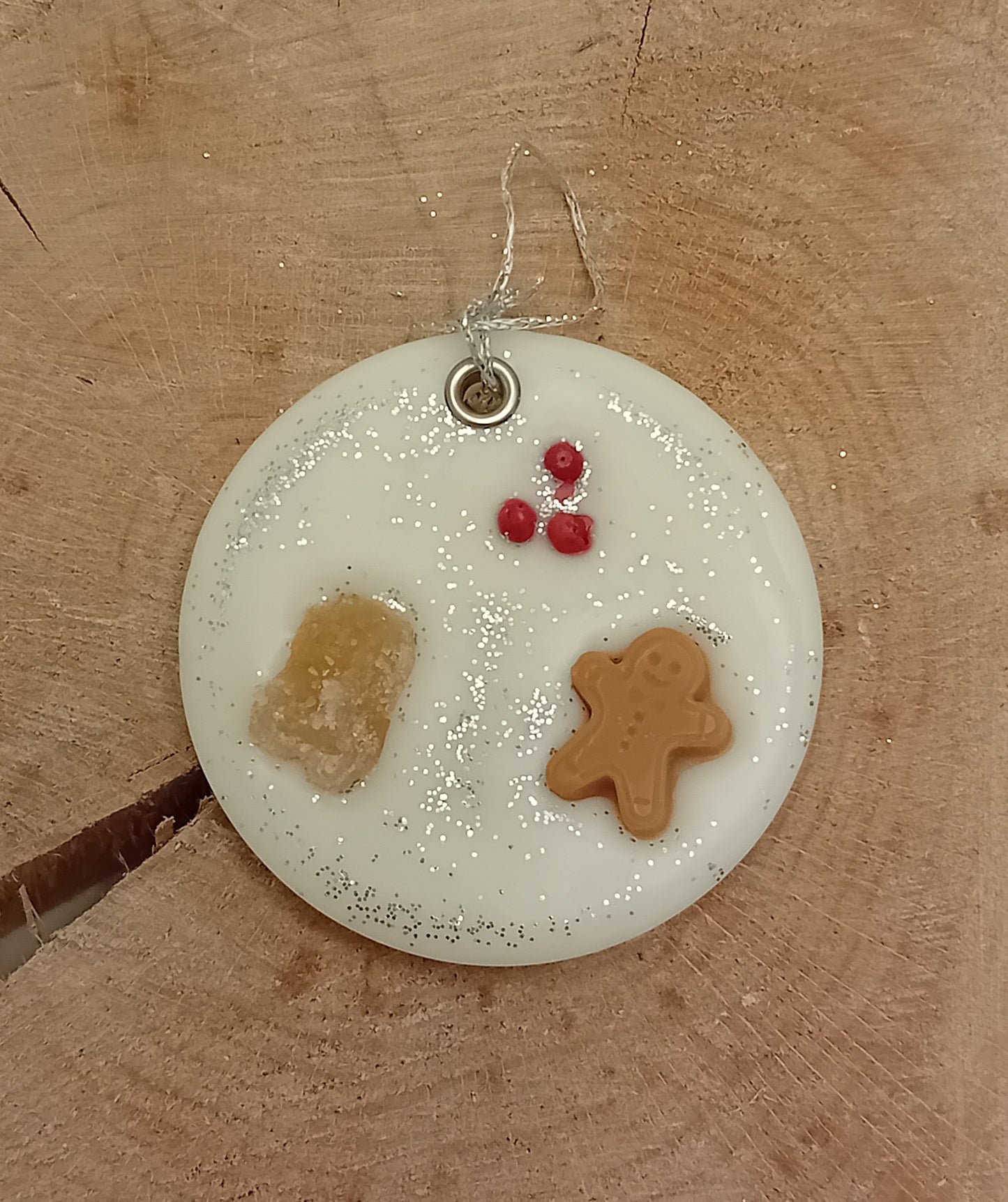 Circular wax sachet decorated with a piece of crystallized ginger, a wax gingerbread man, pink peppercorns and silver glitter with a wooden log slice background.