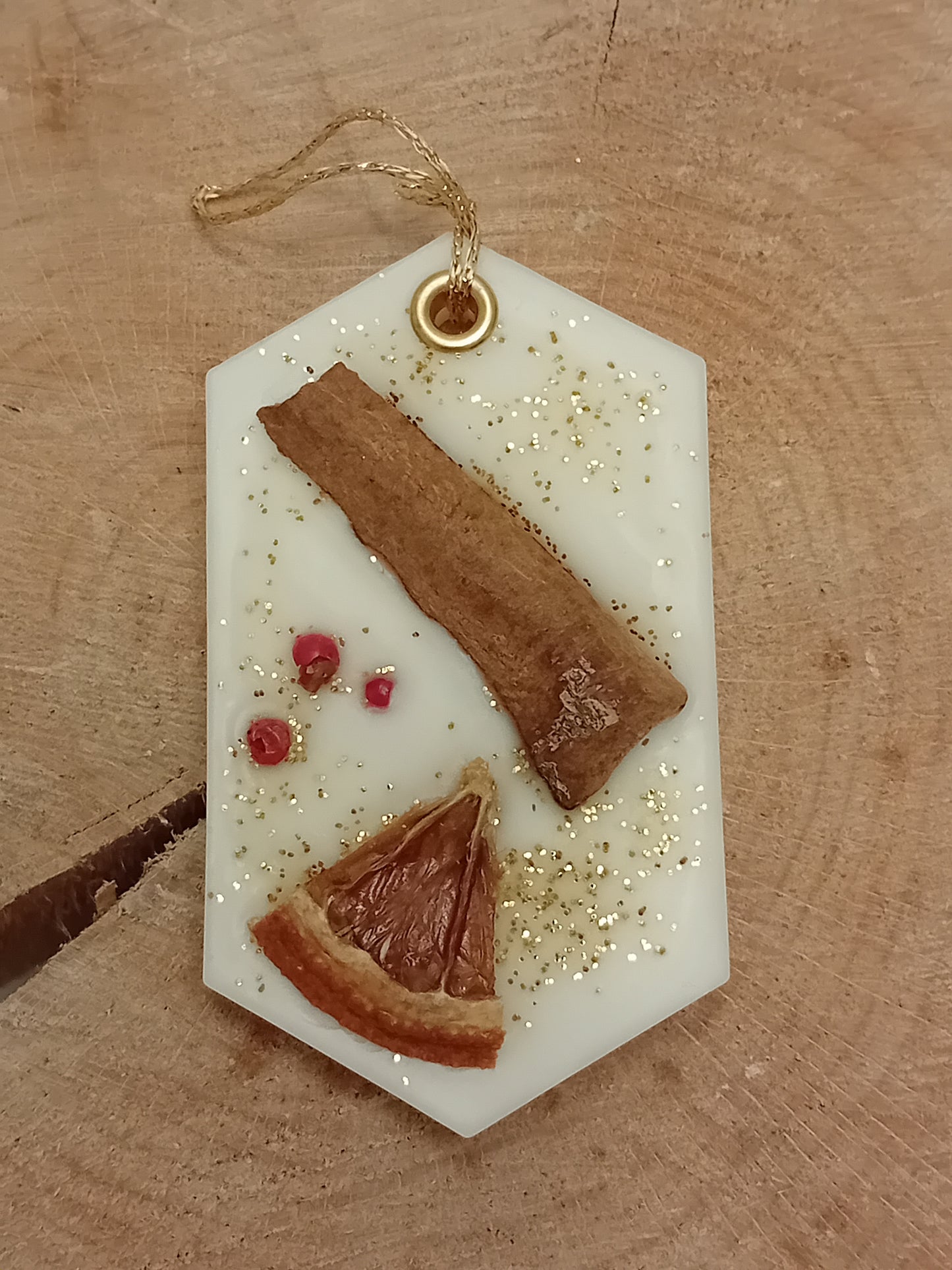 Hexagonal wax sachet decorated with a cinnamon stick, orange slice, pink peppercorns and gold glitter with a wooden log slice background.