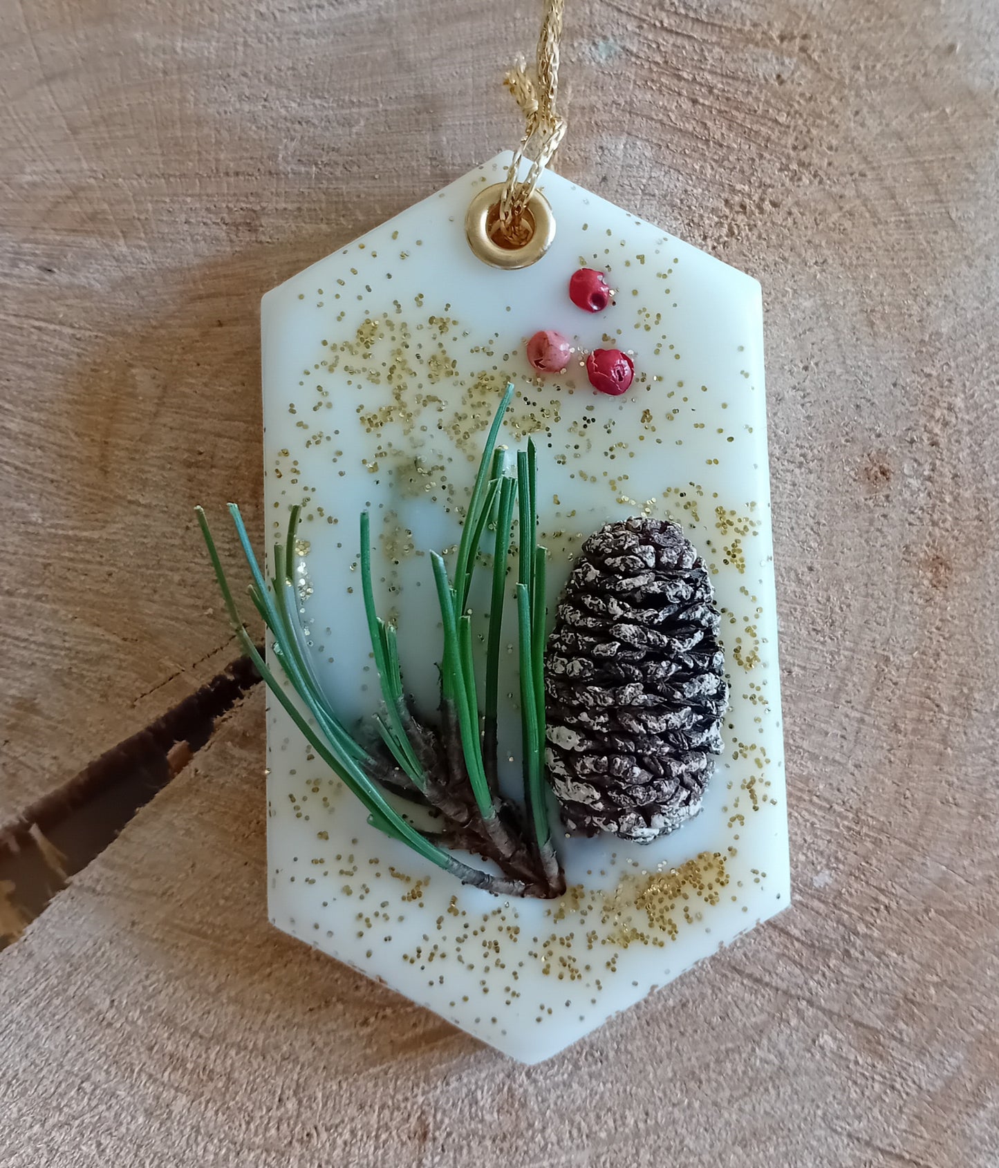 Hexagonal wax sachet decorated with a birch pine cone, pine tree leaves, pink peppercorns and gold glitter with a wooden log slice background.