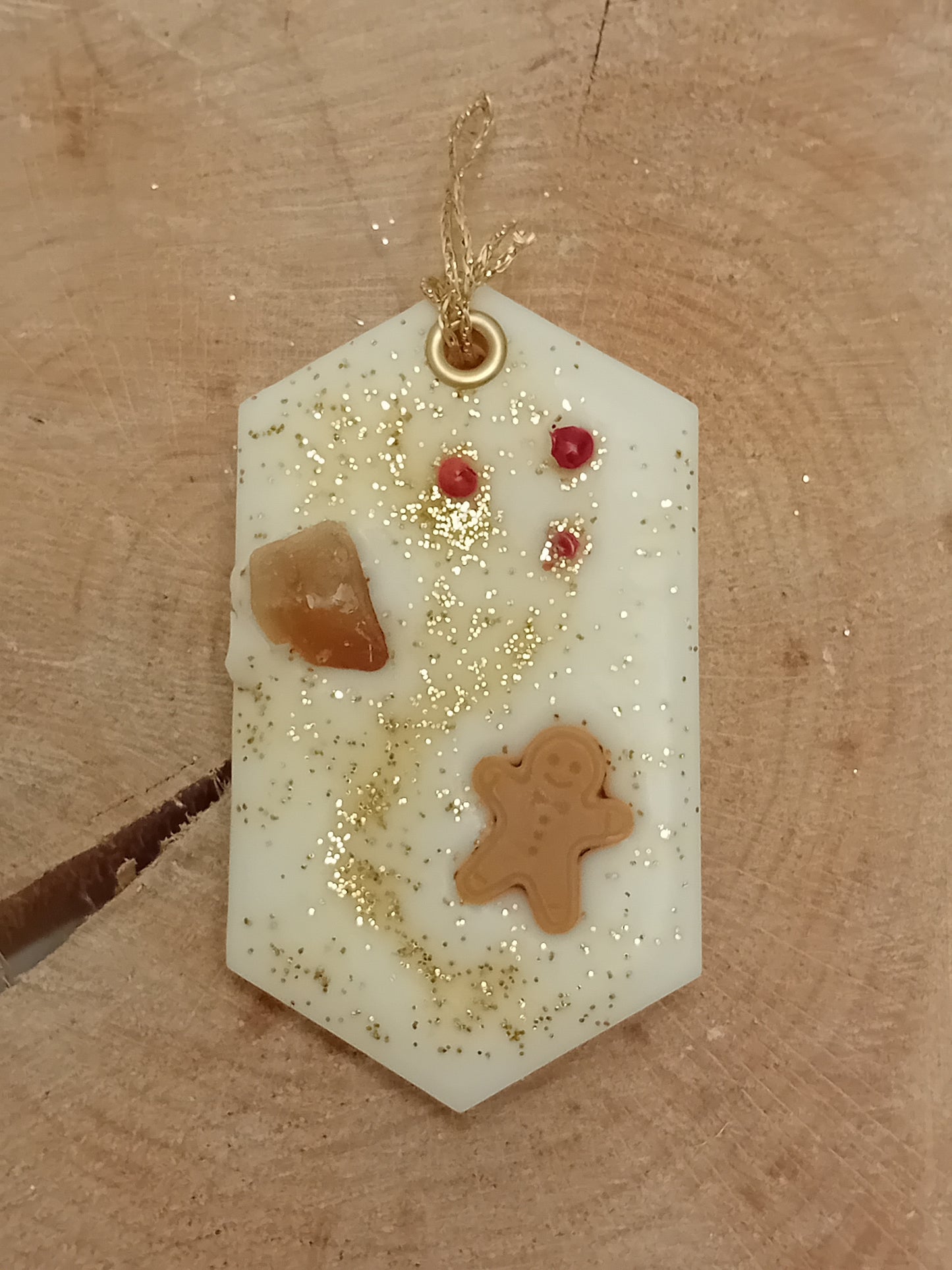 Hexagonal wax sachet decorated with a piece of crystallized ginger, a wax gingerbread man, pink peppercorns and gold glitter with a wooden log slice background.