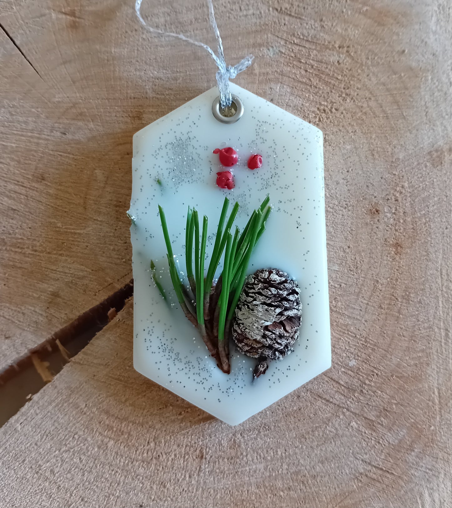 Hexagonal wax sachet decorated with a birch pine cone, pine tree leaves, pink peppercorns and silver glitter with a wooden log slice background.