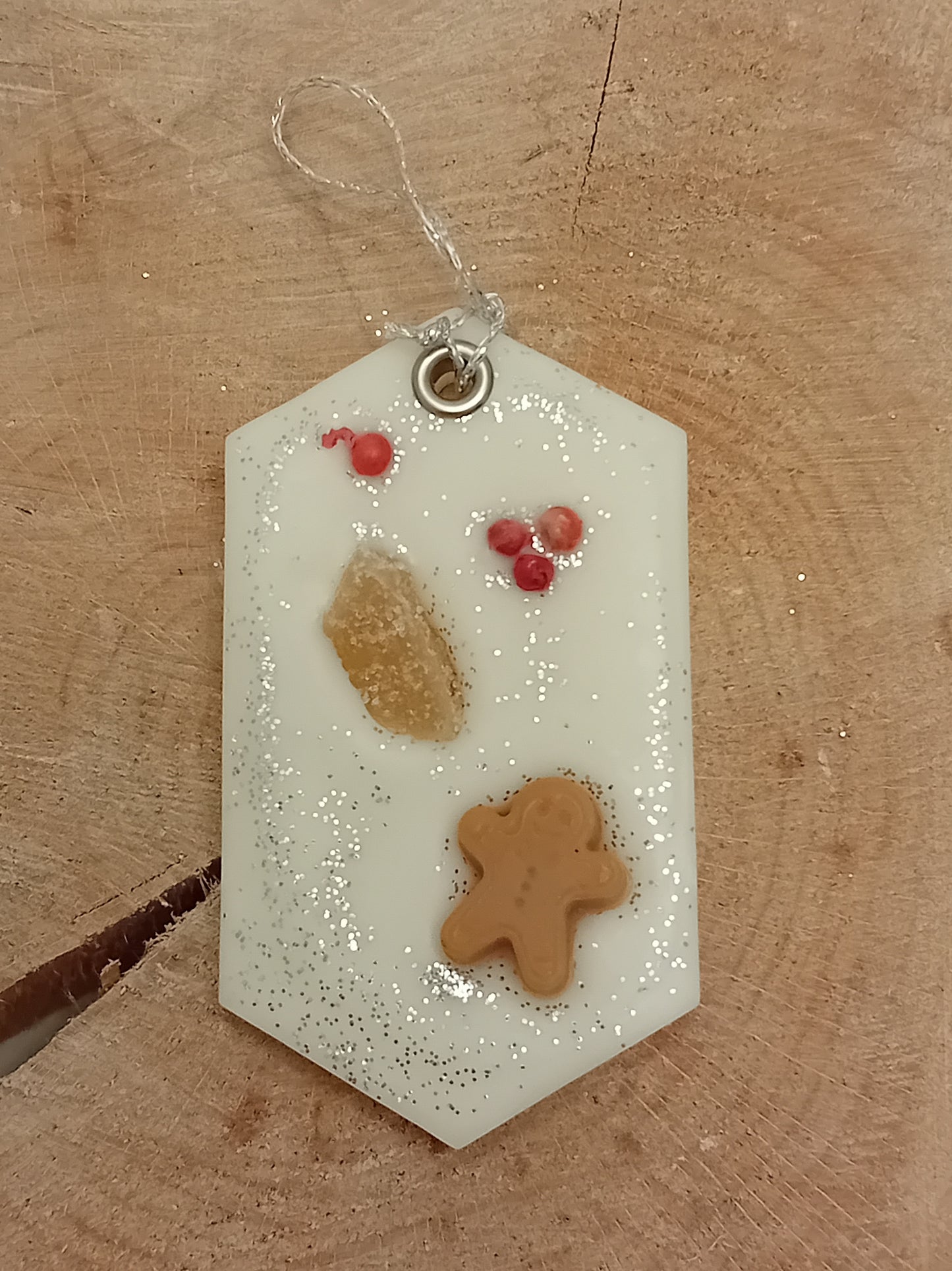 Hexagonal wax sachet decorated with a piece of crystallized ginger, a wax gingerbread man, pink peppercorns and silver glitter with a wooden log slice background.