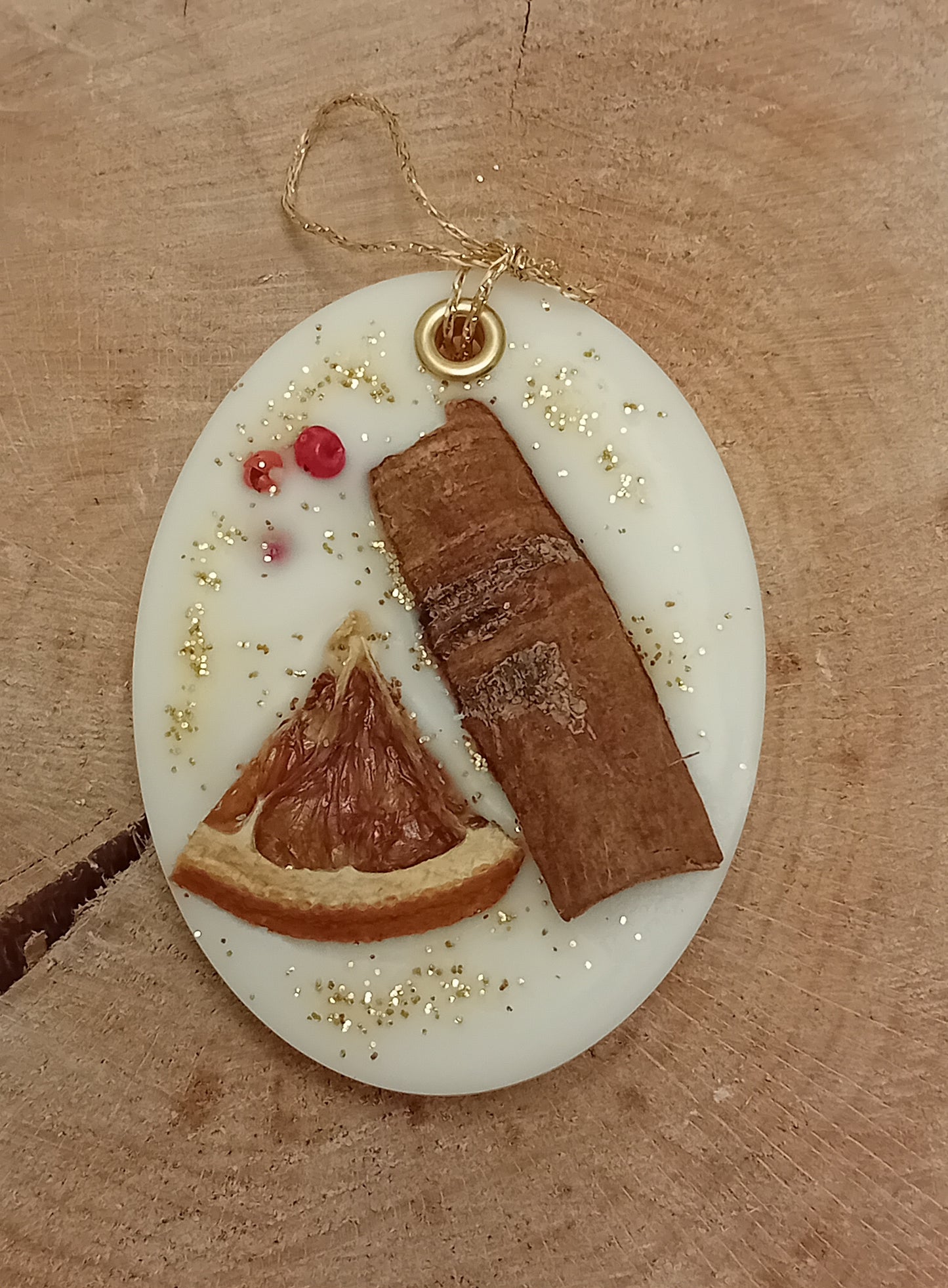 Oval wax sachet decorated with a cinnamon stick, orange slice, pink peppercorns and gold glitter with a wooden log slice background.