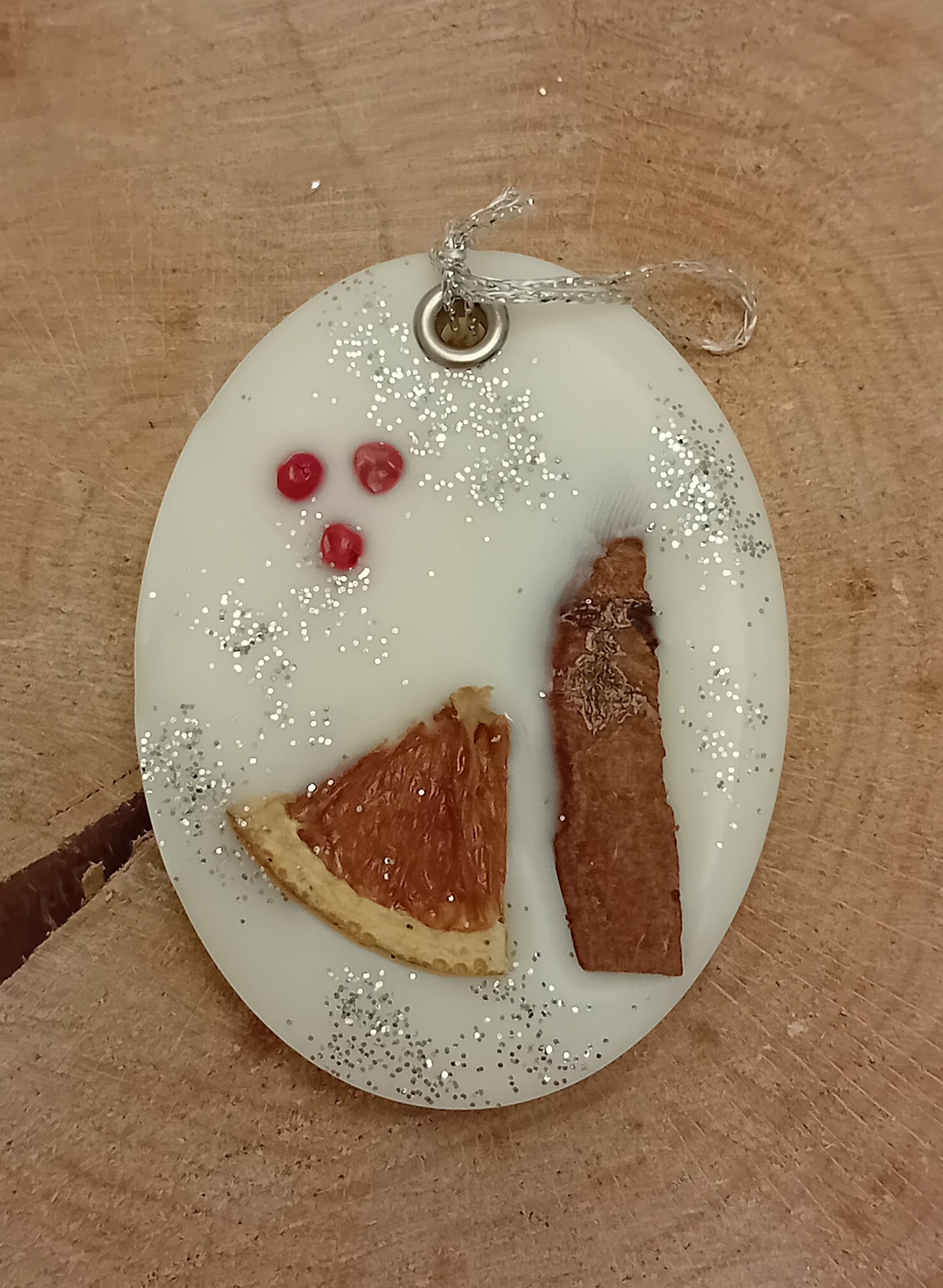 Decorative oval wax sachet with a cinnamon stick, a slice of orange, pink peppercorns and silver glitter with a wooden log slice background.