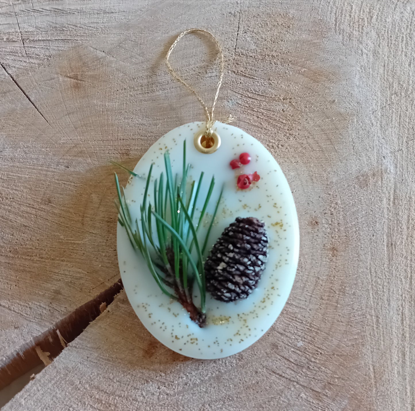 Oval wax sachet decorated with a birch pine cone, pine tree leaves, pink peppercorns and gold glitter with a wooden log slice background.