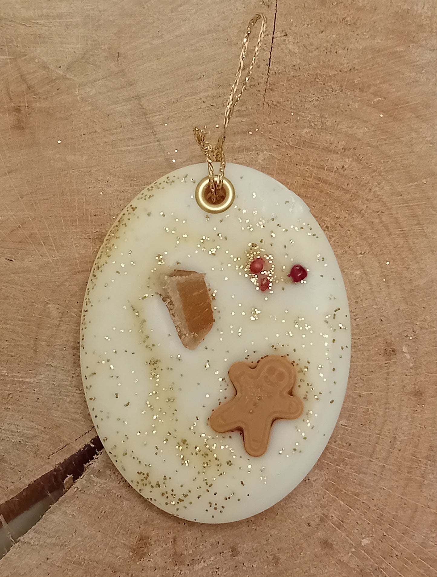 Oval wax sachet decorated with a piece of crystallized ginger, a wax gingerbread man, pink peppercorns and gold glitter with a wooden log slice background.