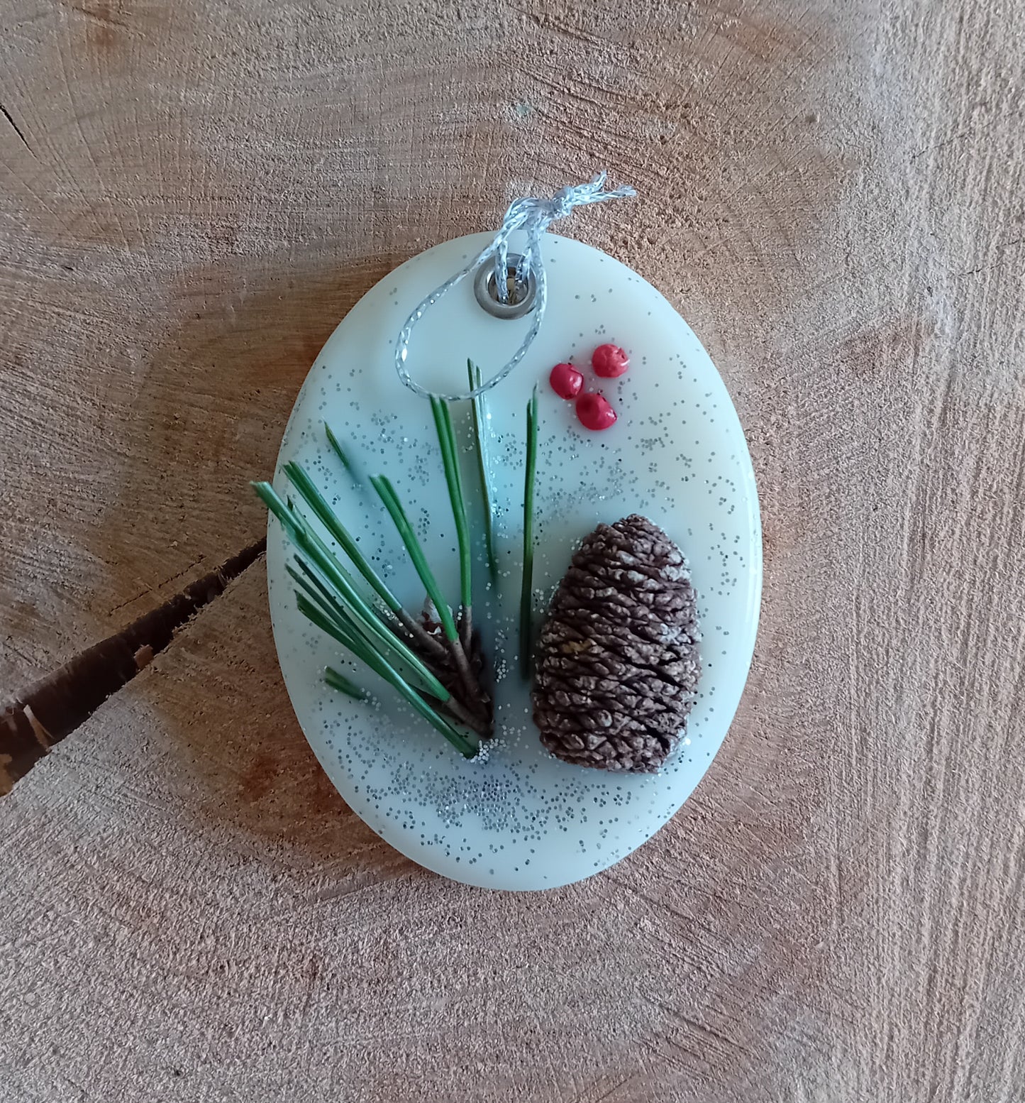 Oval wax sachet decorated with a birch pine cone, pine tree leaves, pink peppercorns and silver glitter with a wooden log slice background.