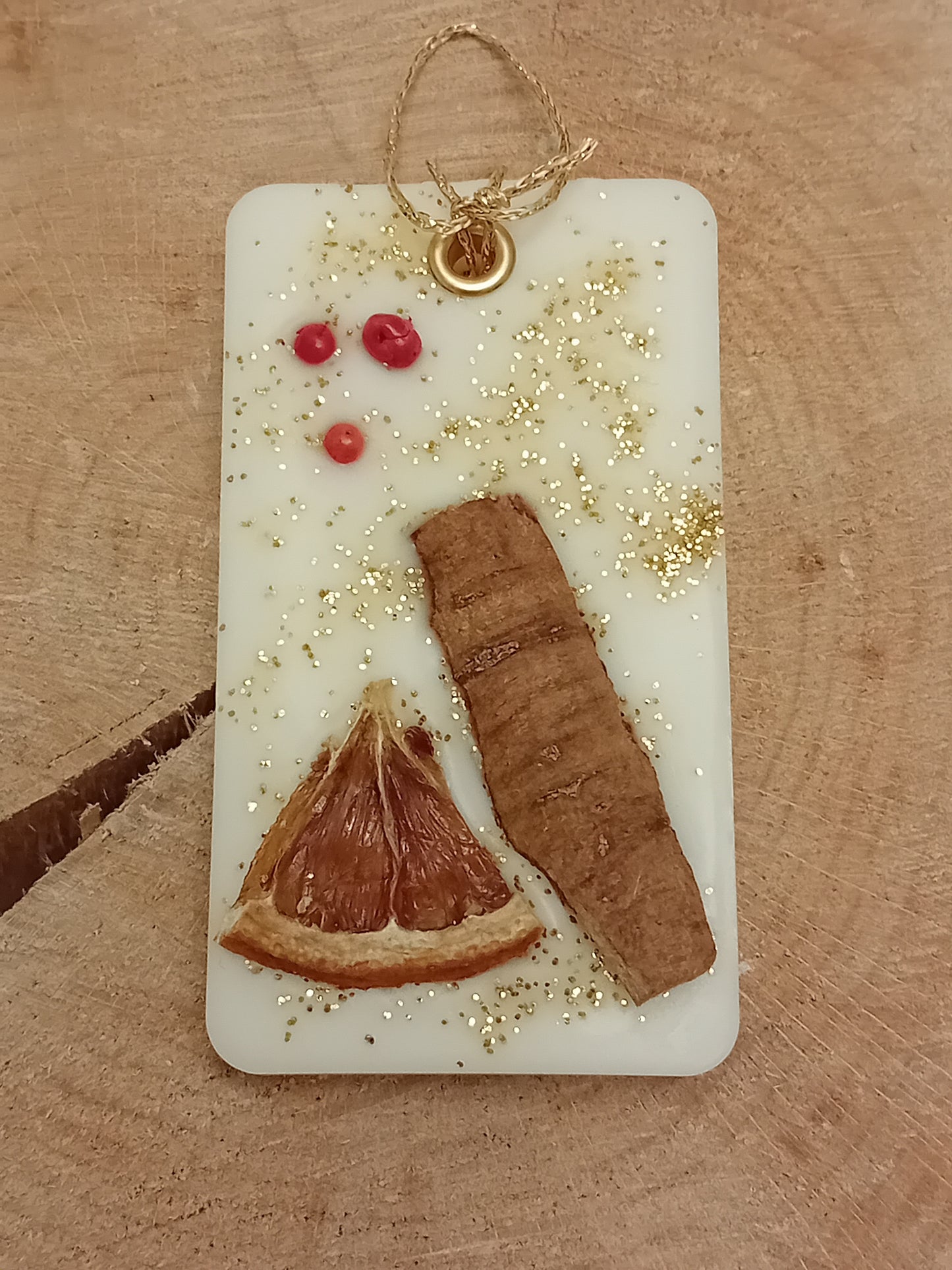 Rectangular wax sachet decorated with a cinnamon stick, orange slice, pink peppercorns and gold glitter on a wooden log slice background.
