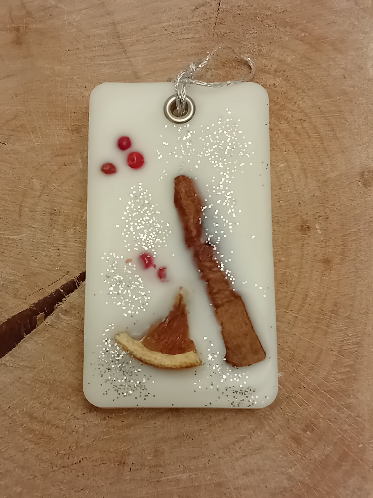 Rectangular wax sachet decorated with a cinnamon stick, orange slice, pink peppercorns and silver glitter on a wooden log slice background.