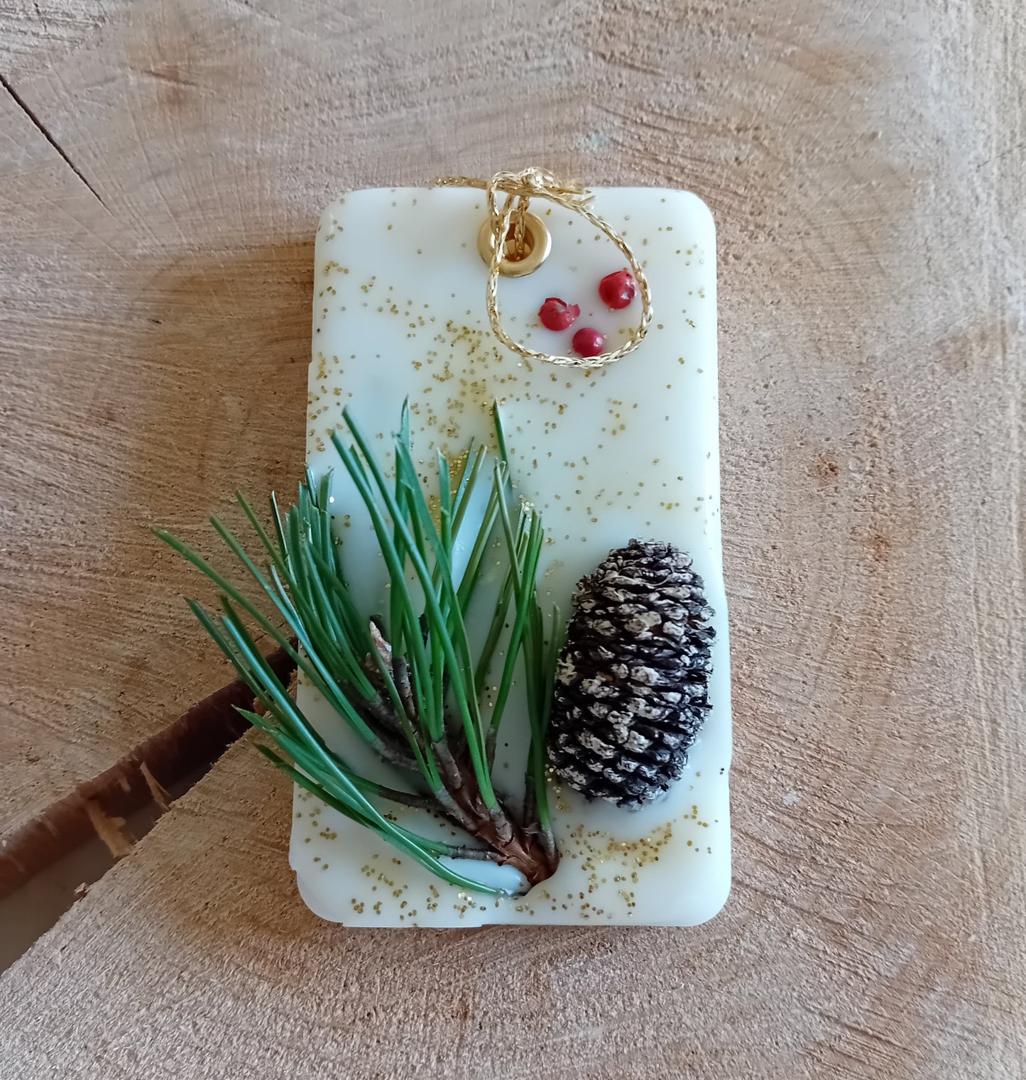 Rectangular wax sachet decorated with a birch pine cone, pine tree leaves, pink peppercorns and gold glitter with a wooden log slice background.