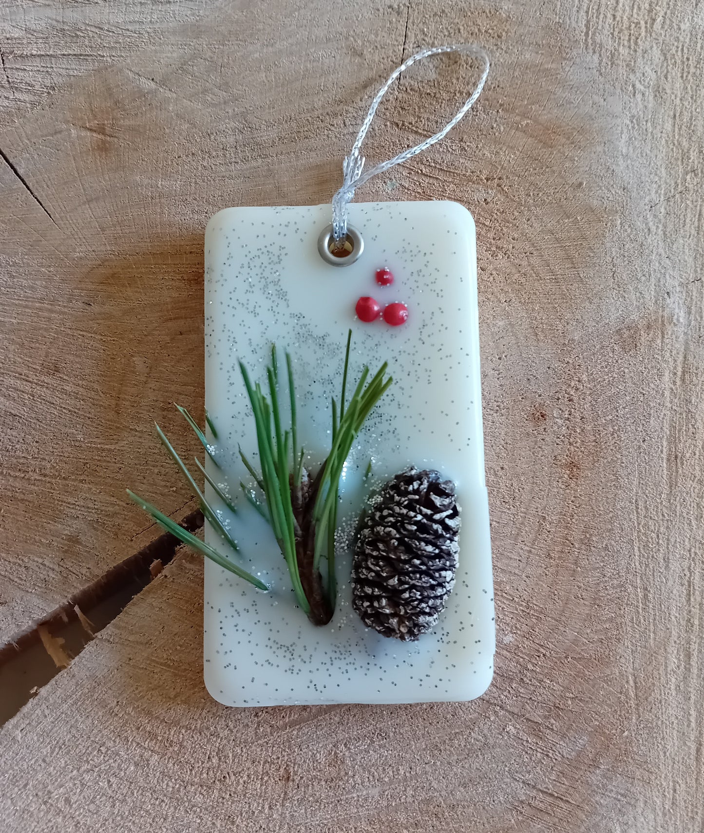 Rectangular wax sachet decorated with a birch pine cone, pine tree leaves, pink peppercorns and silver glitter with a wooden log slice background.