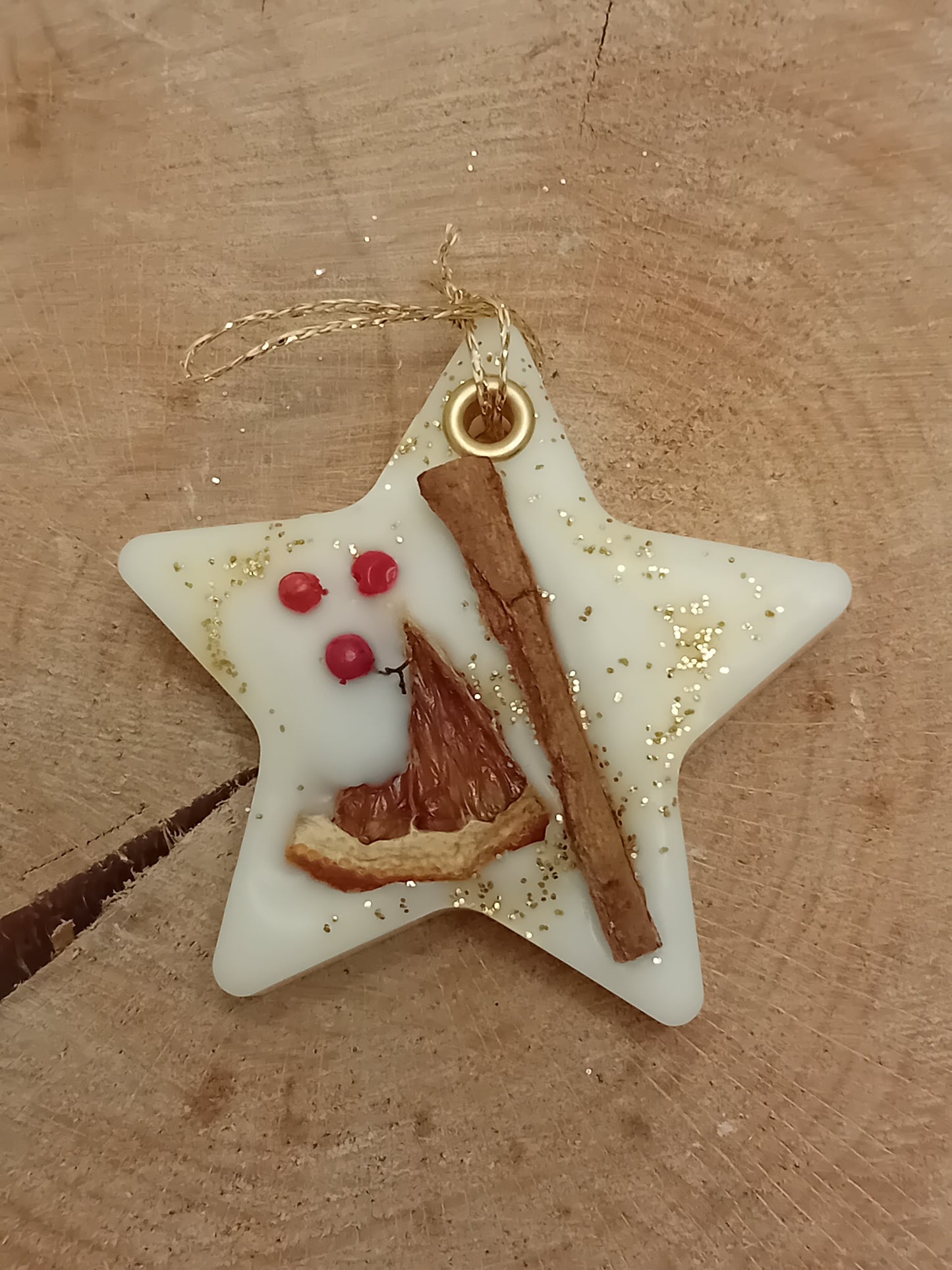 Star-shaped wax sachet decorated with a cinnamon stick, orange slice, pink peppercorns and gold glitter with a wooden log slice background.