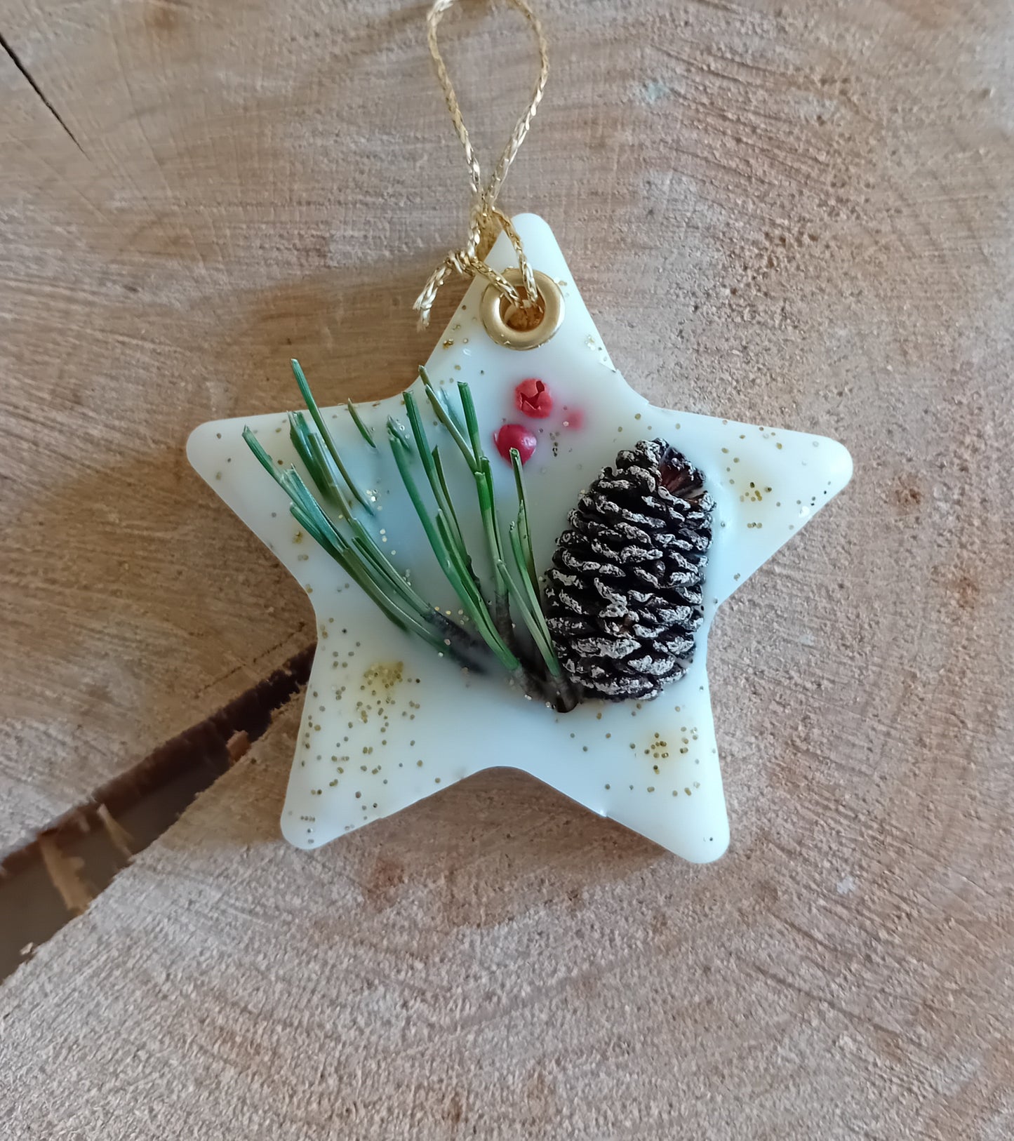 Star-shaped wax sachet decorated with a birch pine cone, pine tree leaves, pink peppercorns and gold glitter with a wooden log slice background.
