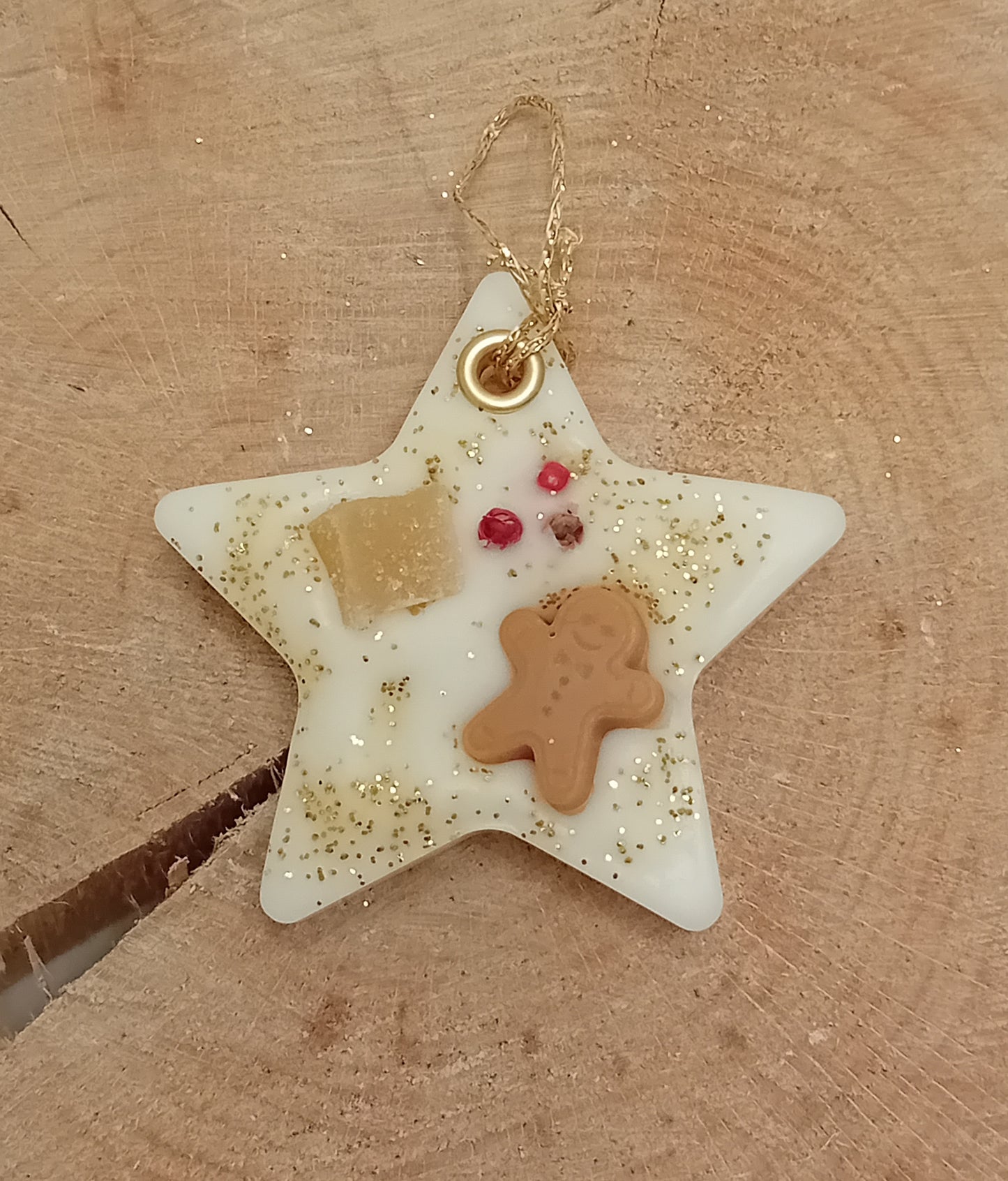 Star-shaped wax sachet decorated with a piece of crystallized ginger, a wax gingerbread man, pink peppercorns and gold glitter with a wooden log slice background.