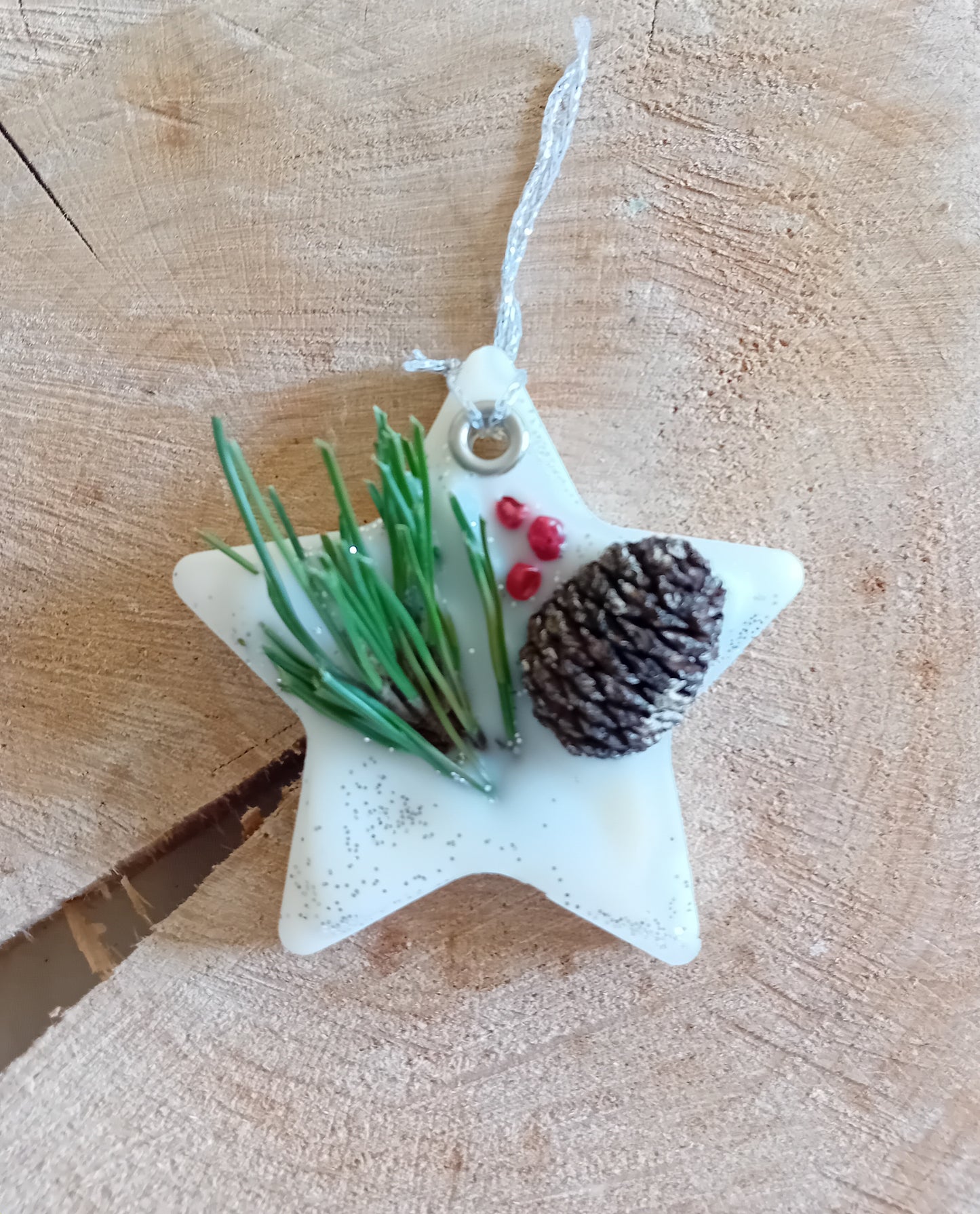 Star-shaped wax sachet decorated with a birch pine cone, pine tree leaves, pink peppercorns and silver glitter with a wooden log slice background.