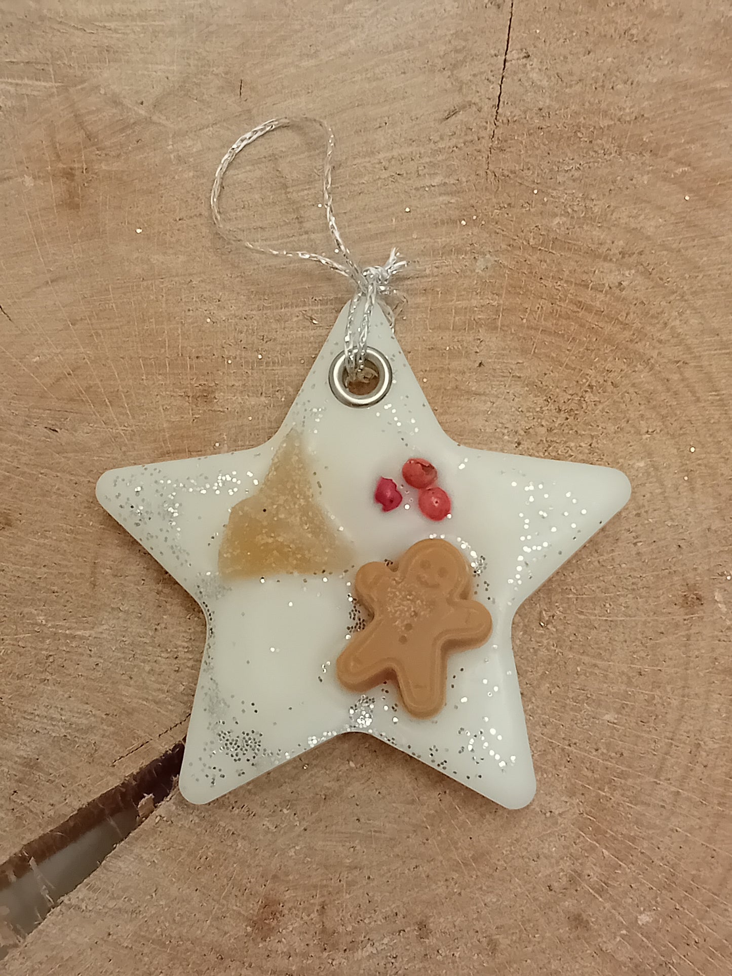 Star-shaped wax sachet decorated with a piece of crystallized ginger, a wax gingerbread man, pink peppercorns and silver glitter with a wooden log slice background.