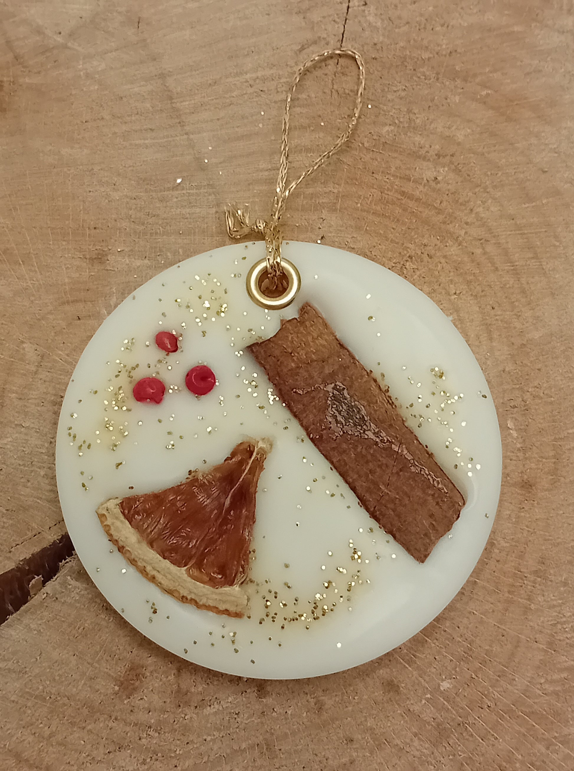 Circular wax sachet decorated with a cinnamon stick, orange slice, pink peppercorns and gold glitter with a wooden log slice background.