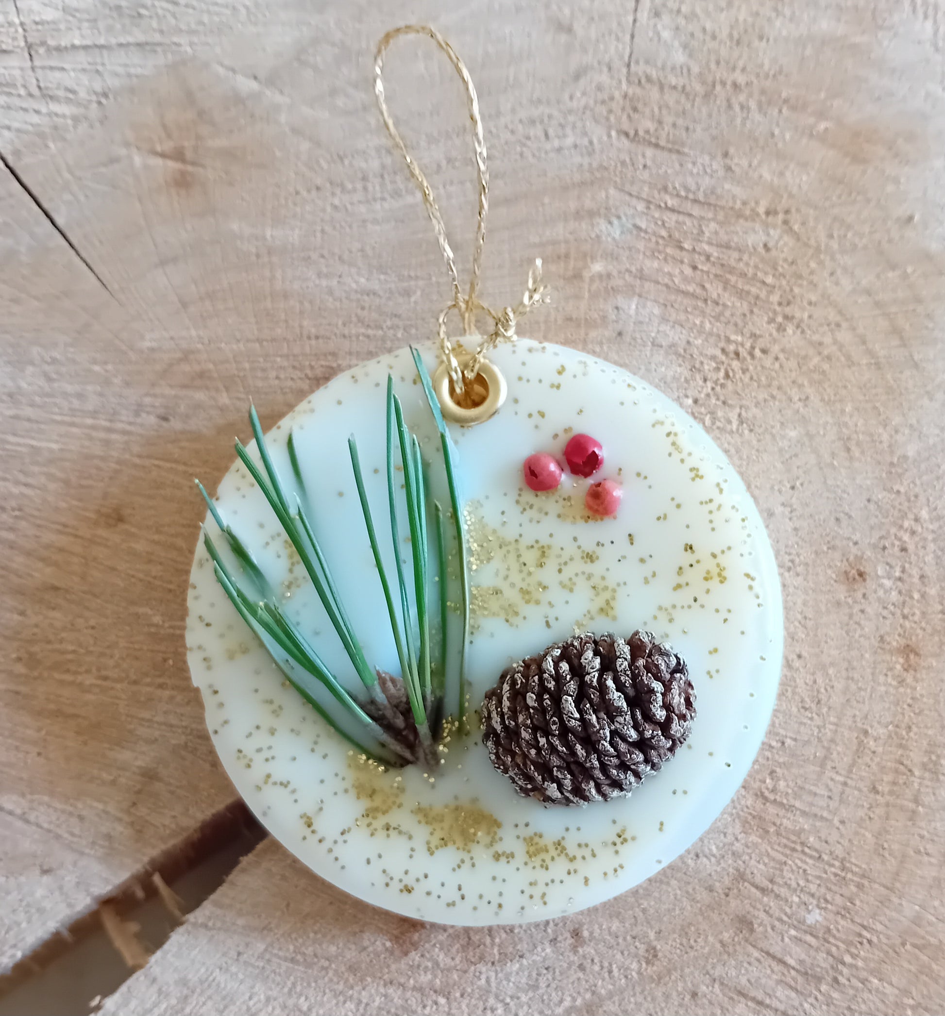 Circular wax sachet decorated with a birch pine cone, pine tree leaves, pink peppercorns and gold glitter with a wooden log slice background.