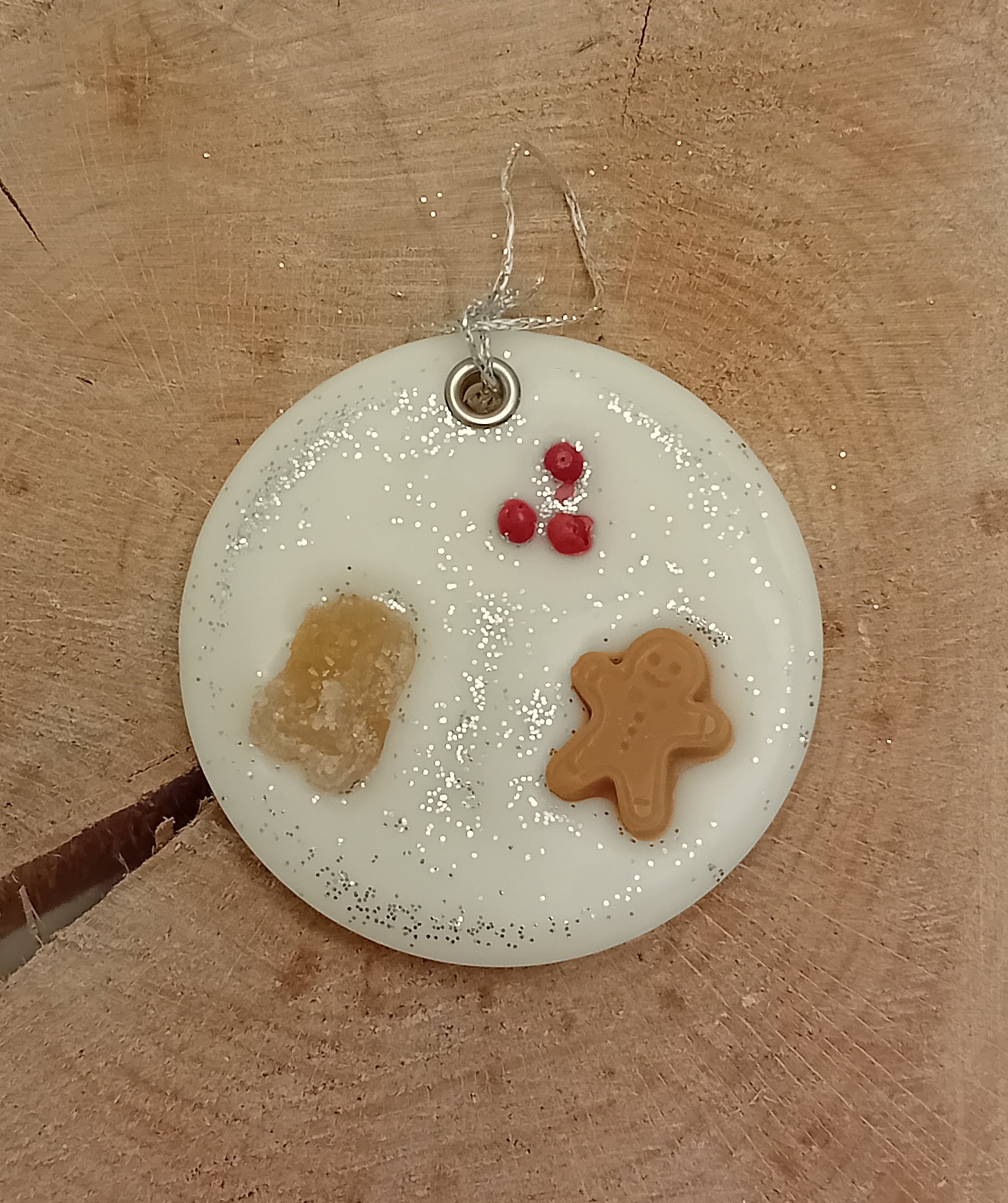 Circular wax sachet decorated with a piece of crystallized ginger, a wax gingerbread man, pink peppercorns and silver glitter with a wooden log slice background.