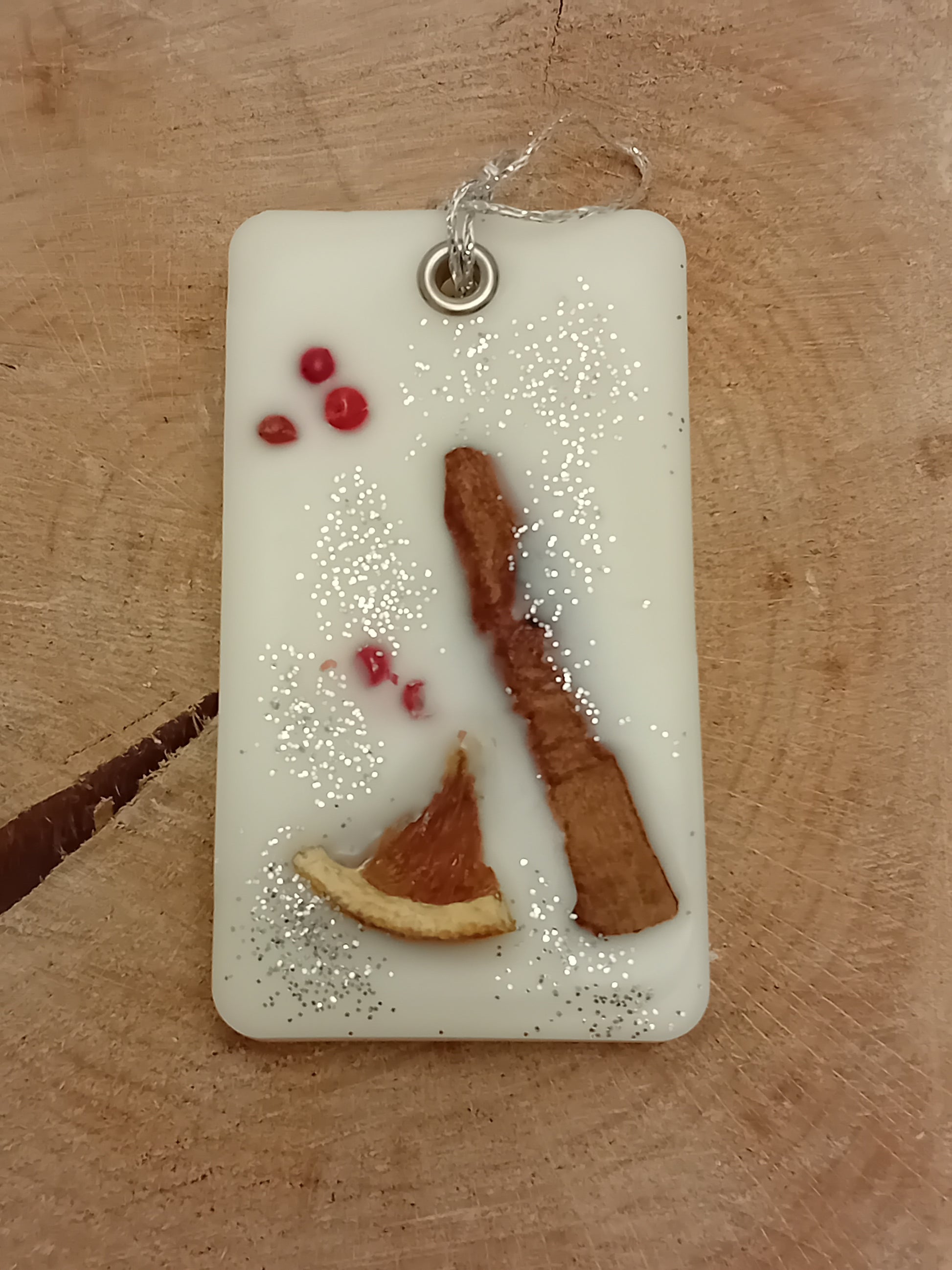 Rectangular wax sachet decorated with a cinnamon stick, orange slice, pink peppercorns and silver glitter on a wooden log slice background.