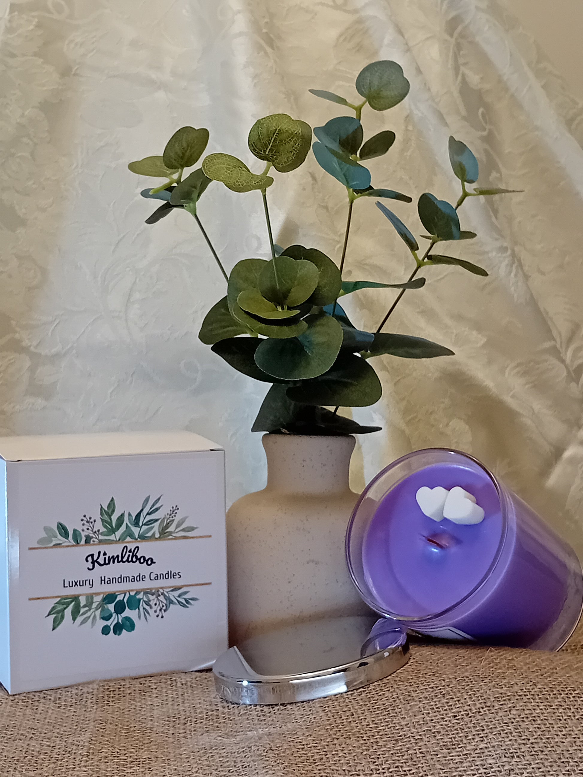 Purple candle in glass jar with 2 mini-heart wax decorations and a silver lid,  next to a white gift box and plant in a vase.